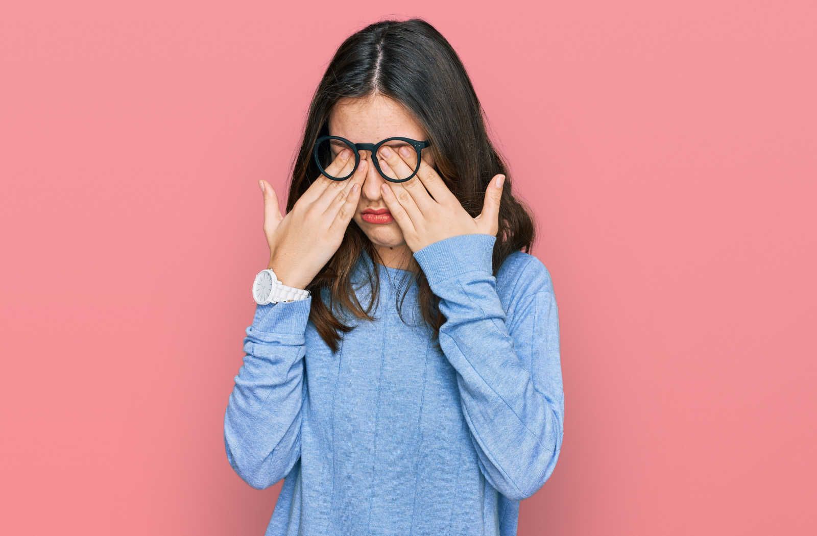 A young girl in a blue sweater, rubbing her eyes due to the discomfort she is feeling as a result of dry eye.