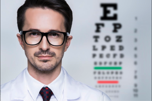 A close up of a male optometrist wearing a pair of glasses with a reading chart in the background.